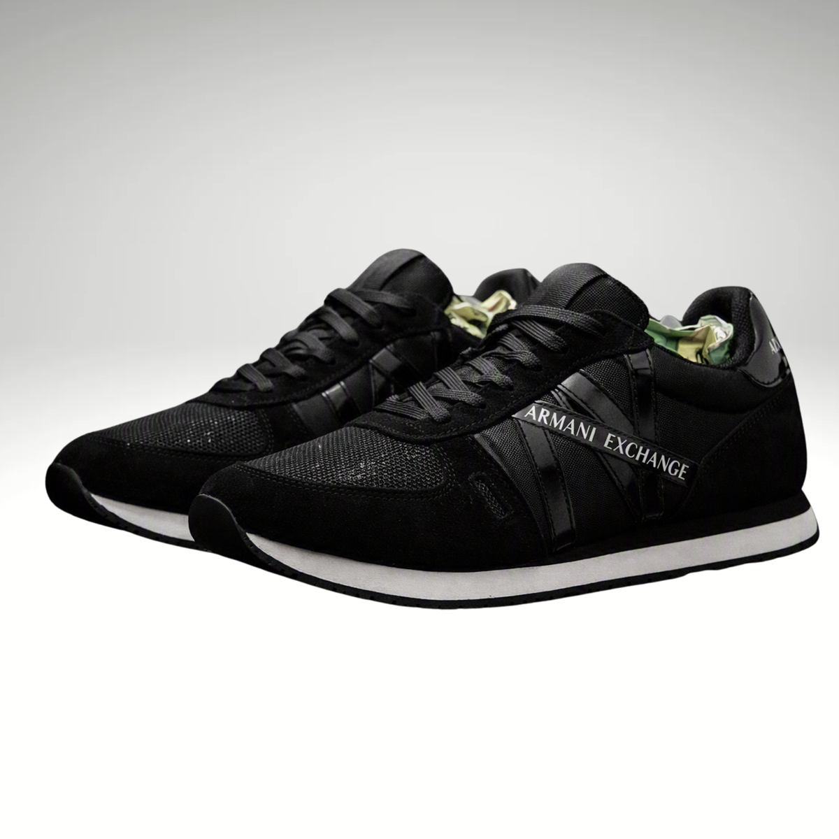 A pair of black Armani Exchange sneakers with black laces, black mesh and suede uppers, and a white midsole, shown against a light gray gradient background.