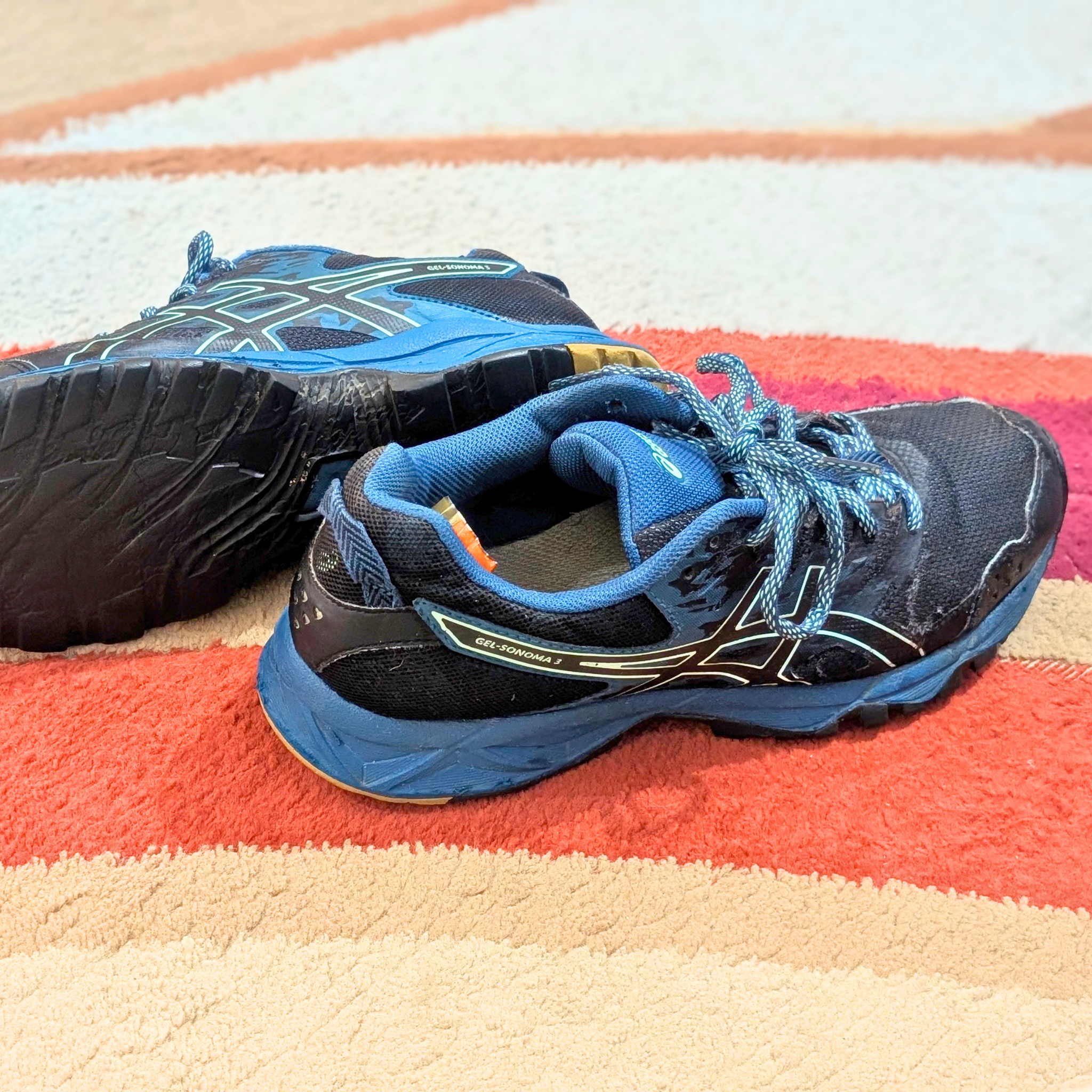 A pair of ASICS running shoes in blue and black, with one shoe showing the side profile and the other showing the outsole, placed on a striped orange, beige, and white rug.