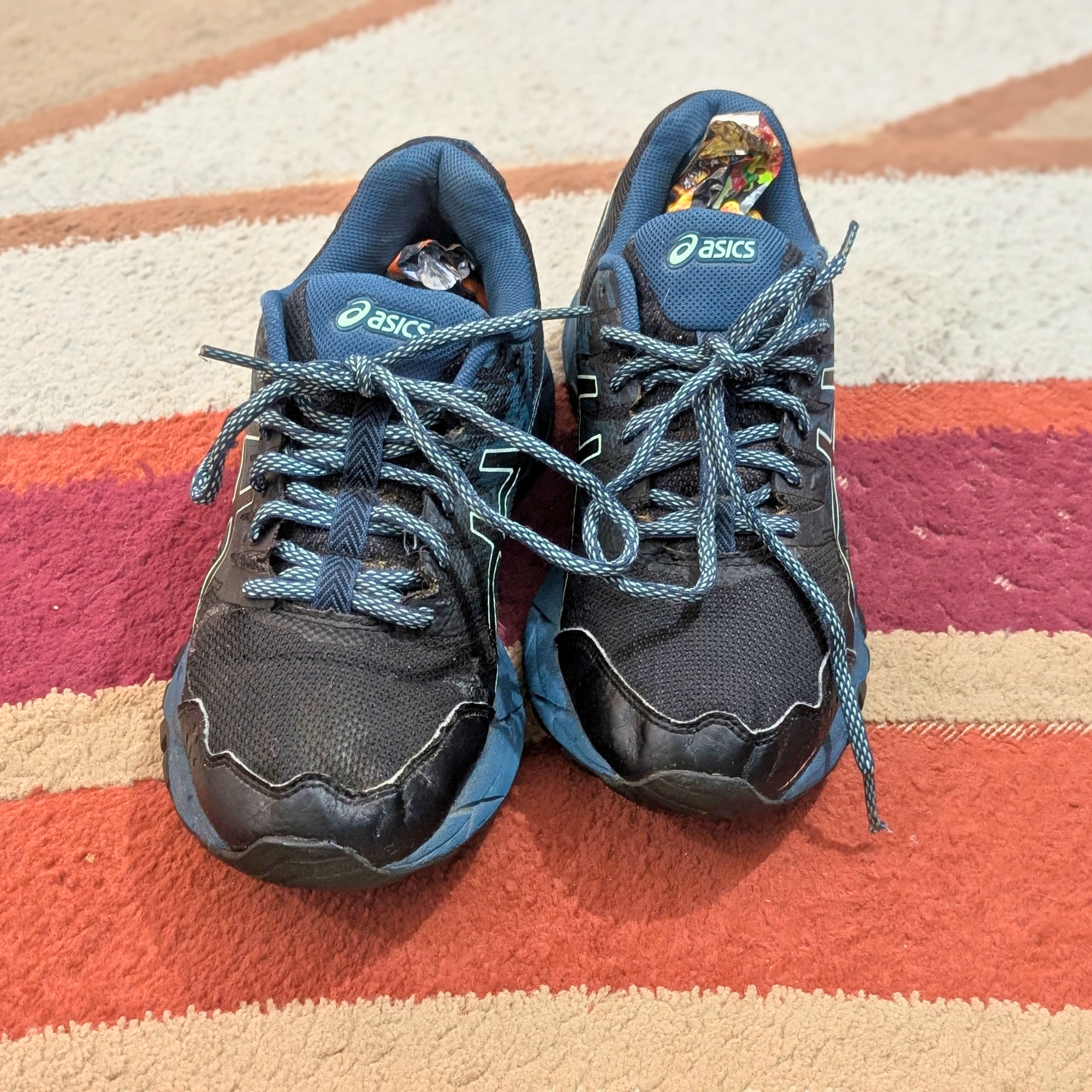 A pair of used Asics running shoes in dark navy and blue, placed on a striped orange, pink, and beige rug. The Asics logos are visible on the tongue and collar.