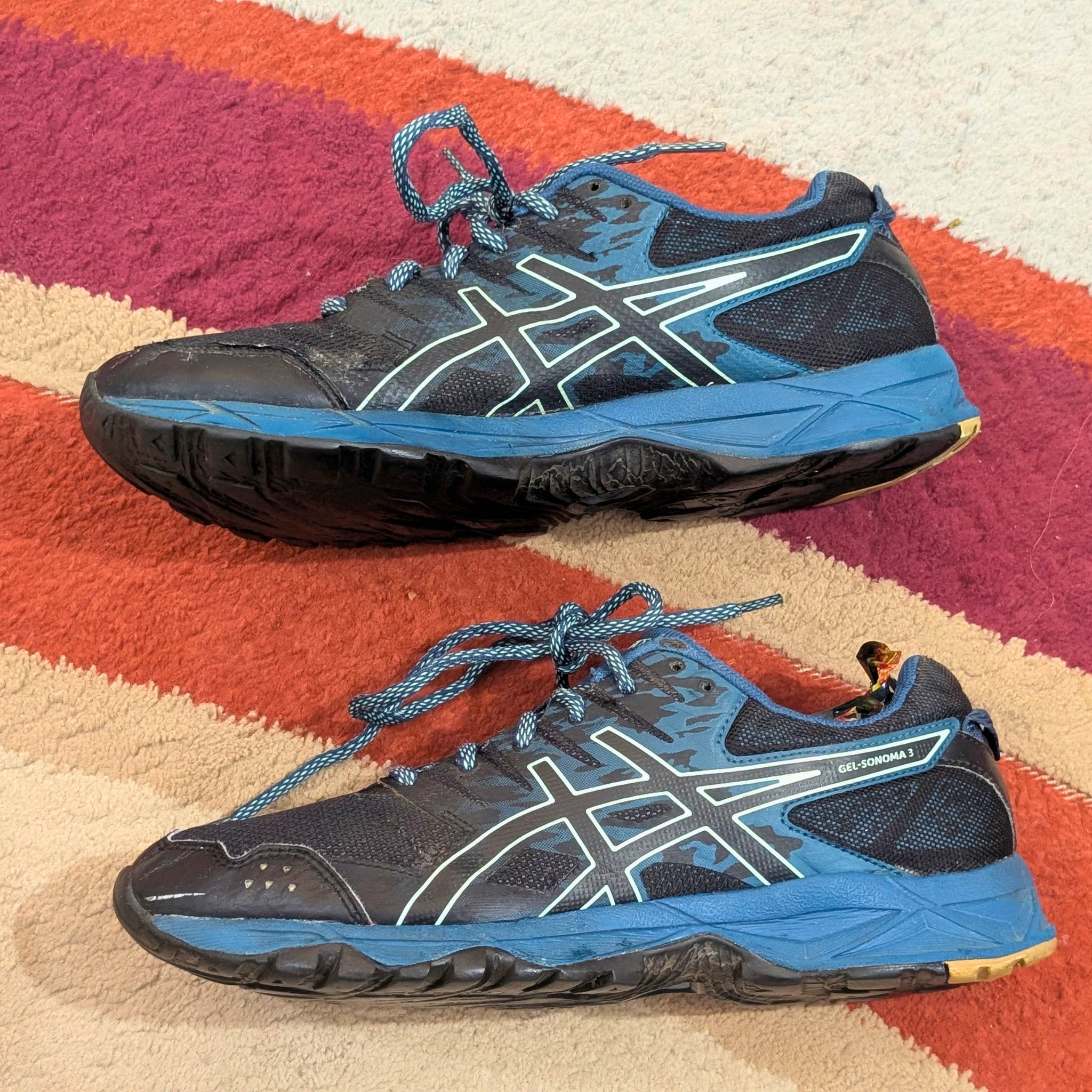 A pair of ASICS GEL-KAYANO 14 running shoes in blue, black, and white, displayed on a striped orange, pink, and beige rug.