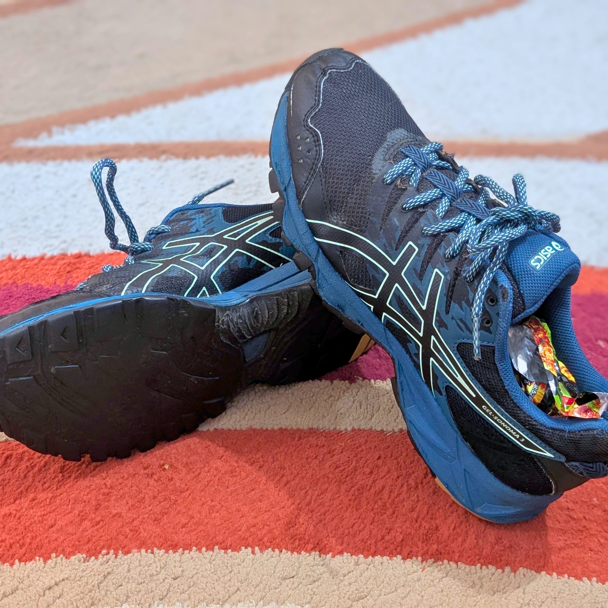 A pair of ASICS running shoes in navy blue and black with light blue laces, resting on a striped red, orange, and beige rug.