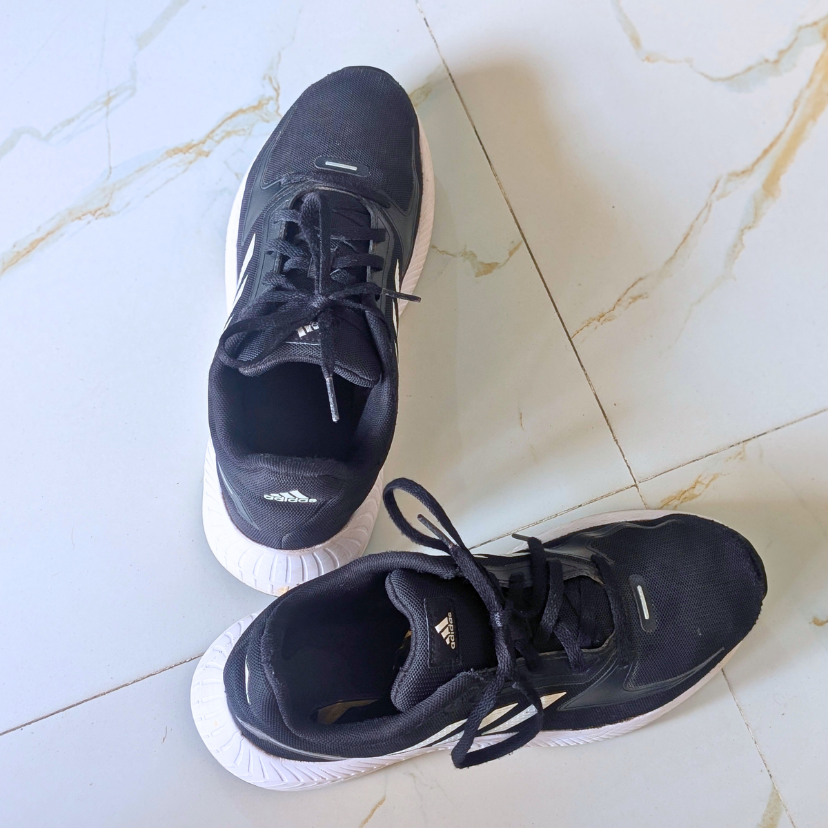 A pair of black Adidas running shoes with white soles, photographed from above on a light marble-tiled floor.