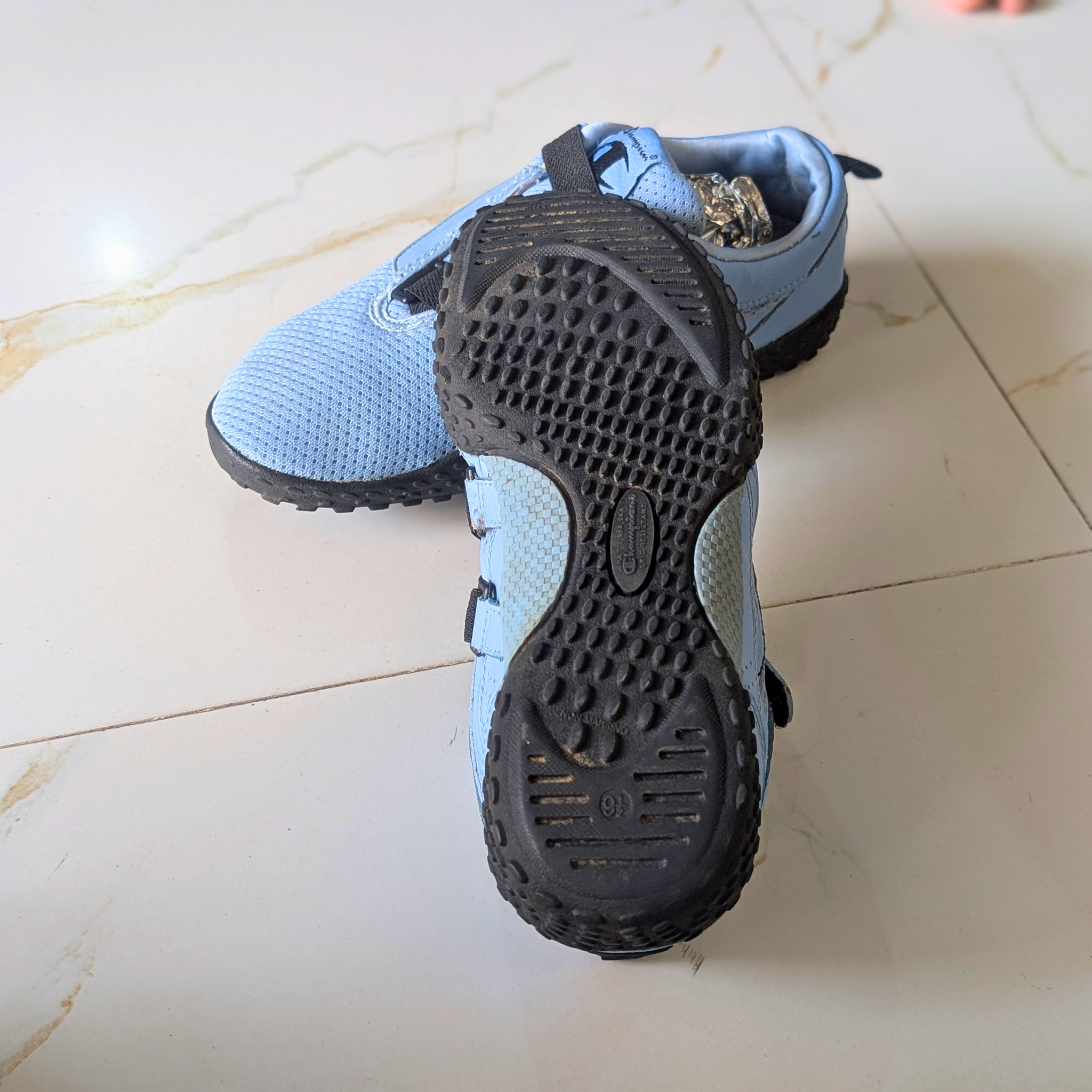 A pair of light blue athletic shoes on a light beige marble floor, with one shoe showing the black textured outsole and the other showing the side profile.