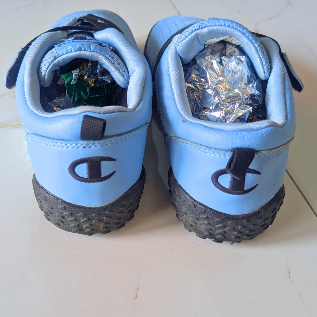 A pair of light blue Champion sneakers viewed from the back, showing the black embroidered 'C' logo and black textured soles. The shoes are resting on a light-colored tiled floor, with crumpled aluminum foil stuffed inside the open back compartments.