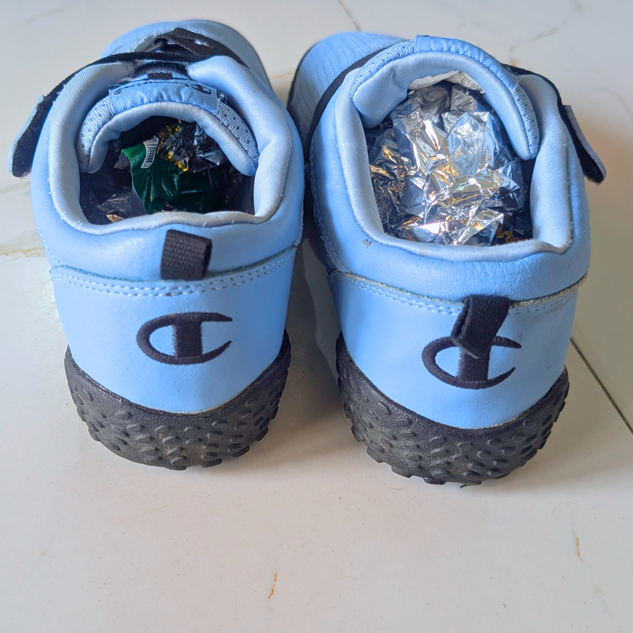 A pair of light blue Champion sneakers viewed from the back, showing the black embroidered 'C' logo and black textured soles. The shoes are resting on a light-colored tiled floor, with crumpled aluminum foil stuffed inside the open back compartments.