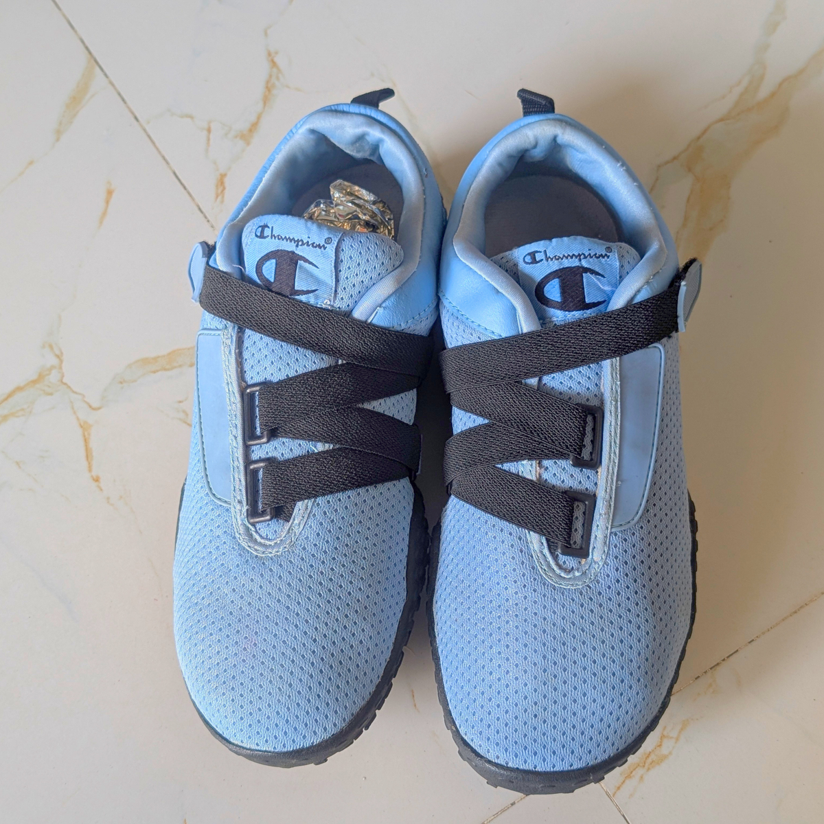 A pair of light blue Champion slip-on shoes with black elastic straps, laid flat on a beige marble-patterned floor.