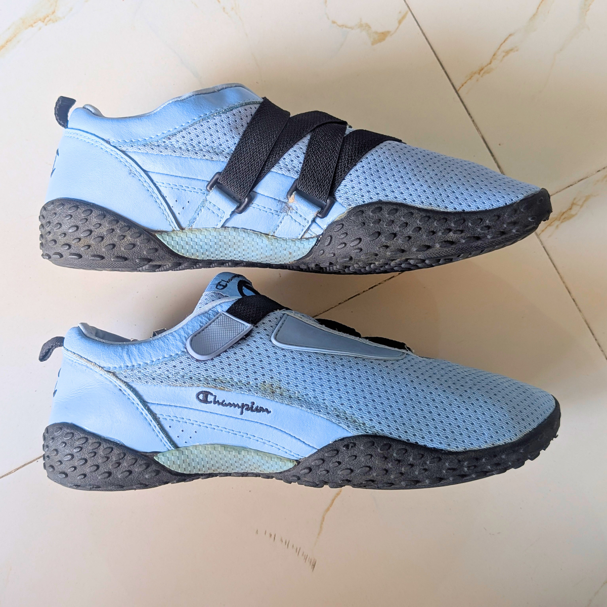 A pair of Champion athletic sneakers in light blue and black, displayed on a beige tiled floor.