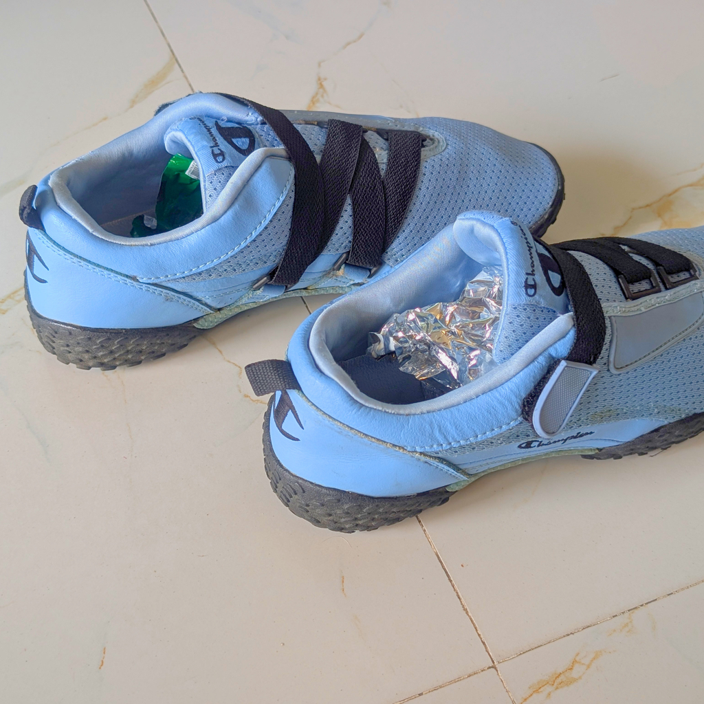 A pair of light blue athletic shoes with black laces and black soles, placed on a beige tiled floor. One shoe is shown from the side and the other from the top, with crumpled aluminum foil inside the opening of the right shoe.