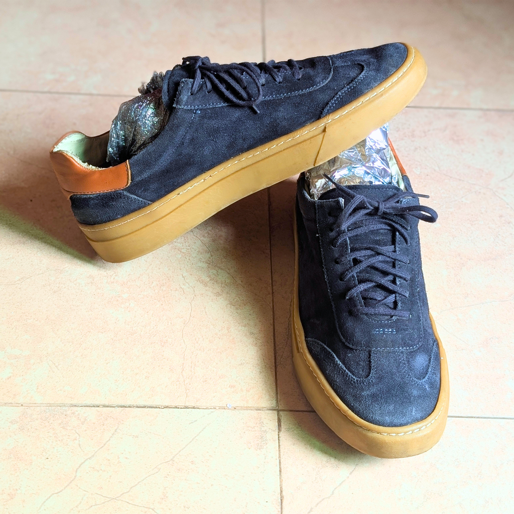 A pair of low-top suede sneakers in dark navy blue with tan rubber soles and a small brown leather heel tab, photographed on a beige tiled floor.