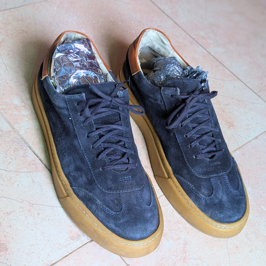 A pair of dark navy blue suede low-top sneakers with tan rubber soles, displayed on a light beige tiled floor. The shoes are shown with their interiors open, revealing silver crumpled paper inserts.