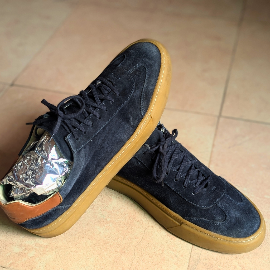 A pair of low-top suede sneakers in dark navy blue with gum brown soles, displayed on a beige tiled floor. One shoe is positioned upright while the other lies on its side, showing a silver crumpled foil insert inside the heel area.