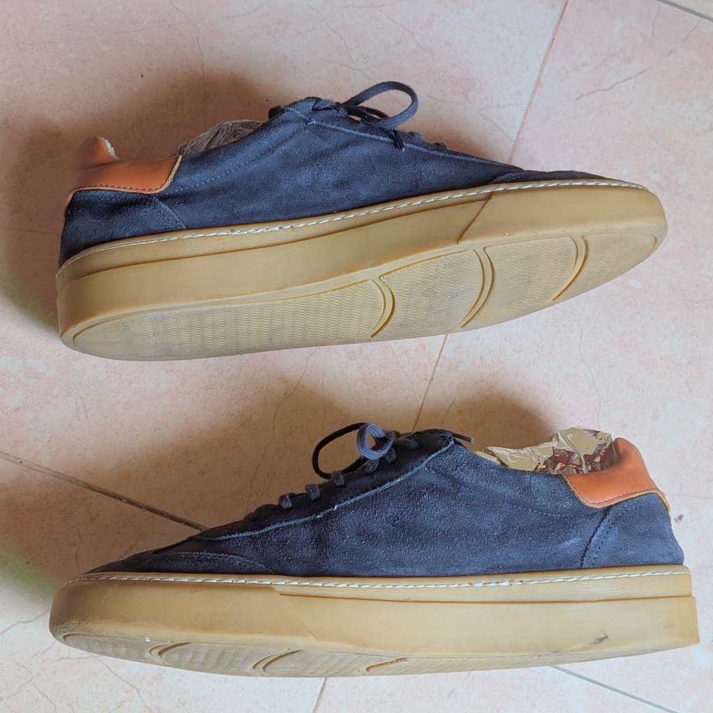 A pair of low-top casual sneakers with navy blue suede uppers, tan leather heel tabs, and thick beige rubber soles, shown from top and side views on a tiled floor.