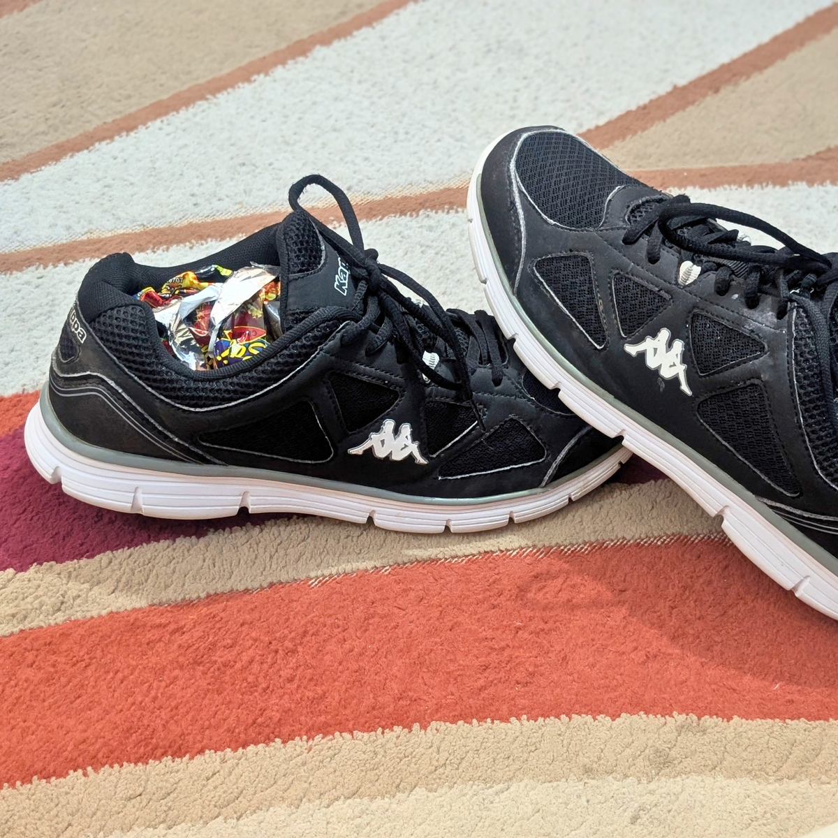 A pair of black Kappa athletic sneakers with white soles, resting on a striped beige, rust, and brown textured rug.