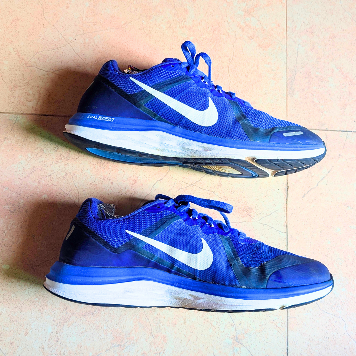 A pair of Nike running shoes in royal blue and white, displayed on a light beige tiled floor.