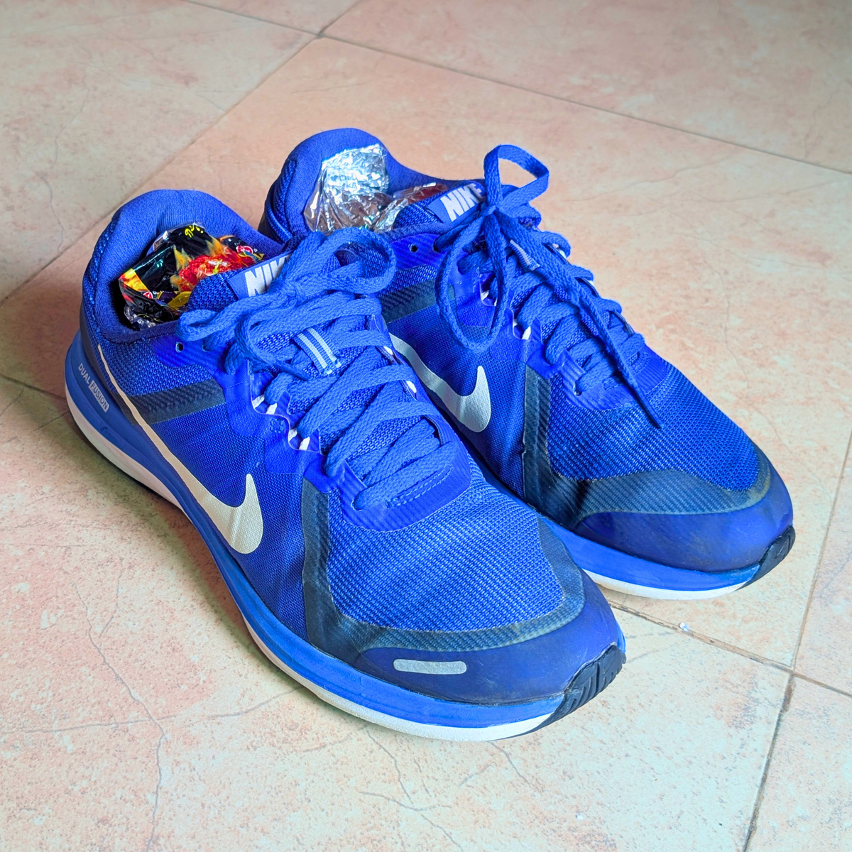 A pair of Nike running shoes in royal blue with white swooshes, placed on a light beige tiled floor.