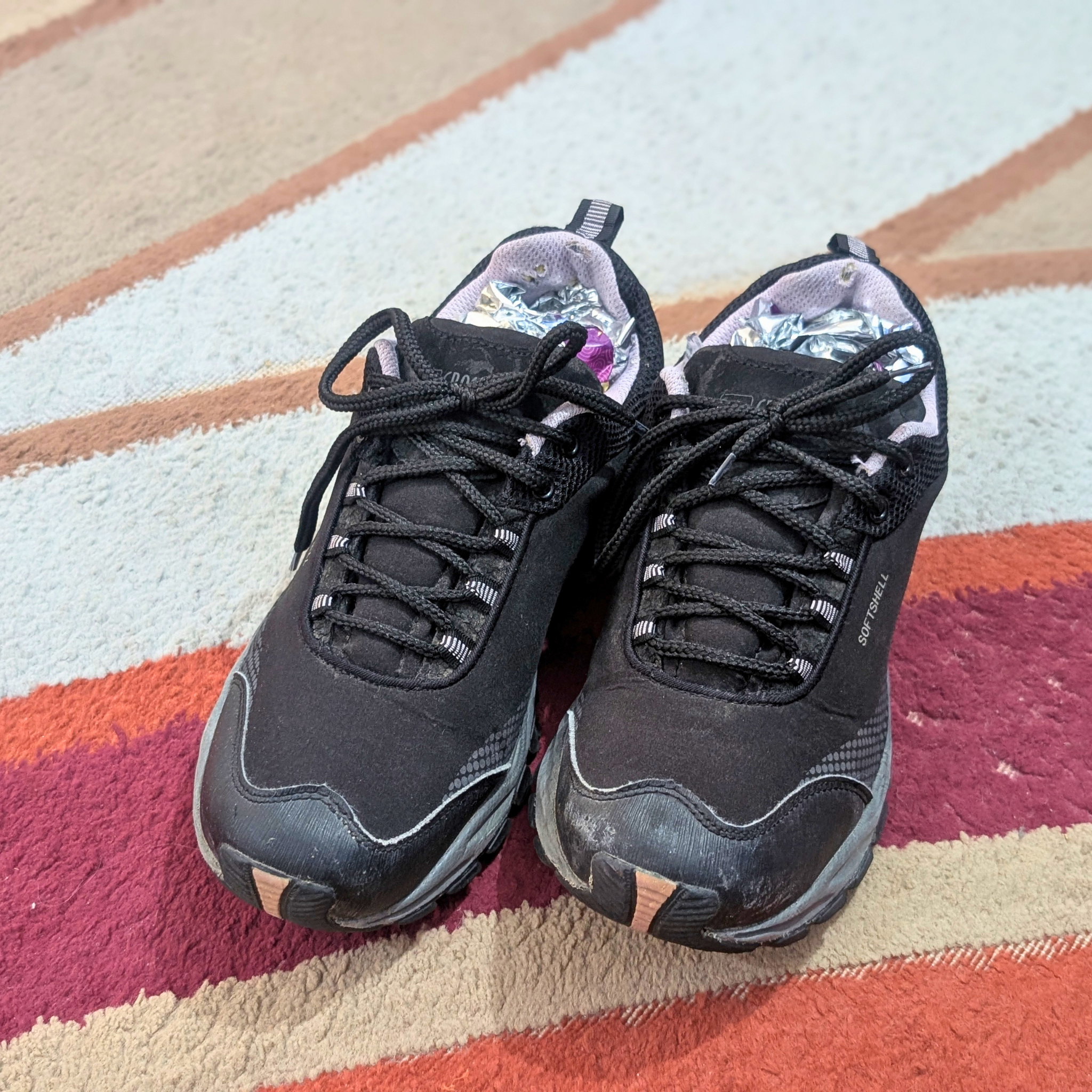 A pair of black athletic trail running shoes with black laces, resting on a striped carpet with beige, orange, and red patterns.